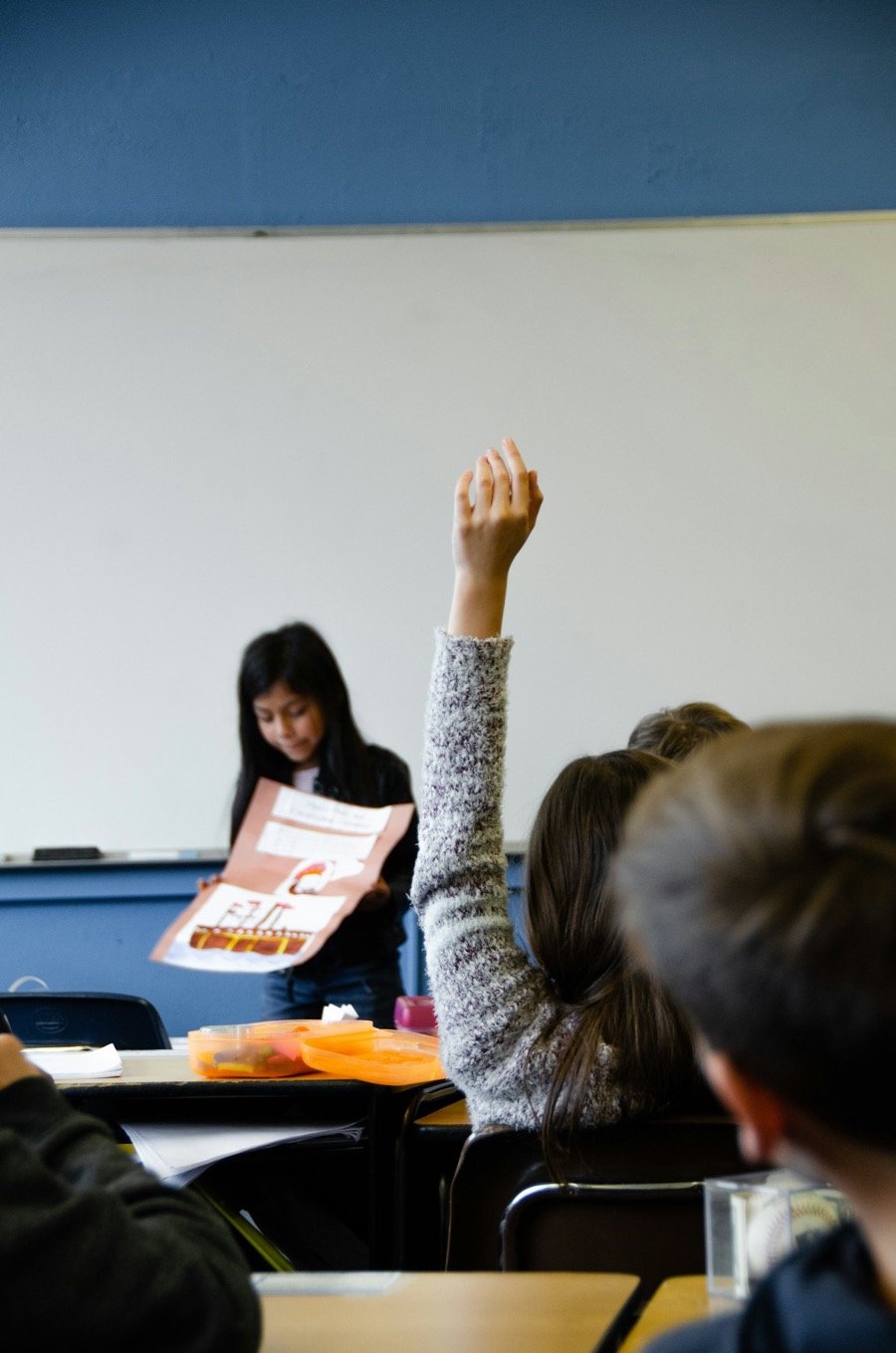 A pupil presents her work to the class while a classmate raises a hand to ask a question