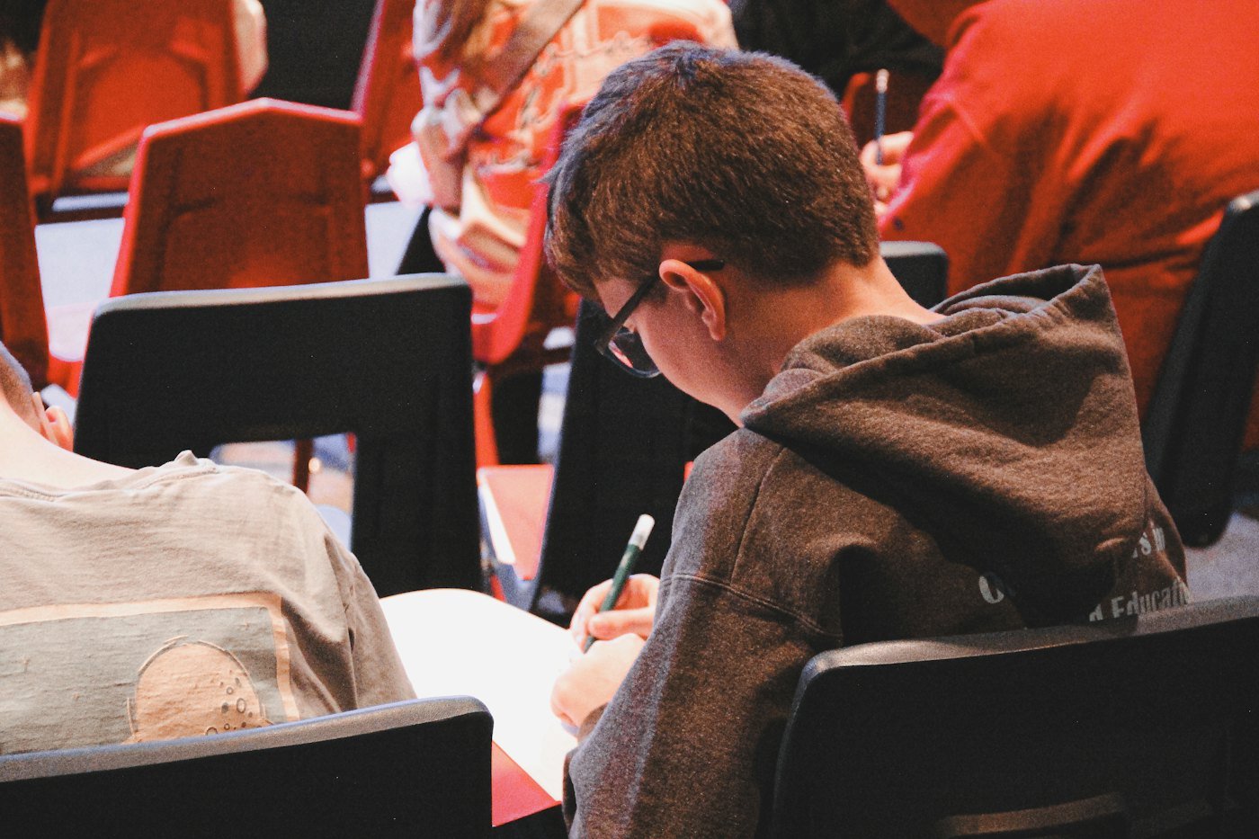 A student in a hall of red seats writes notes on a sheet of paper